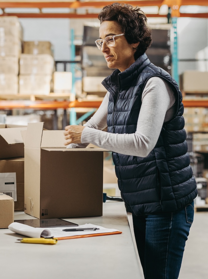 Swagger employee is unboxing product and doing real-time inventory on a table. We see other Swagger branded merch items in boxes on the table and in the background, there is a view of Swagger’s storage facility with organized boxes on palettes.